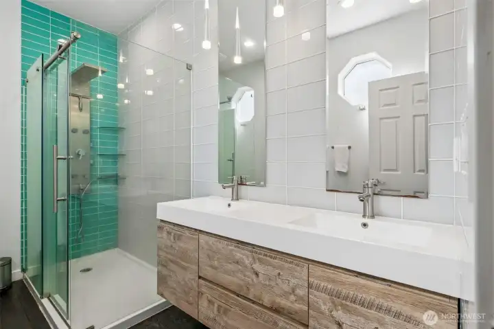 The shower is fully dialed in with floor-to-ceiling tile, a low-threshold entry, and a rain-style fixture that shifts the whole experience into spa territory. The tile brings in a rich, elevated green tone that adds depth without overpowering the space. Across from it, a floating vanity anchors the room with clean lines, dual sinks, and extra-tall beveled mirrors that amplify both light and scale. The full wall tile backsplash carries that same energy across the room, while the octagon windows punch in architectural interest and natural light.