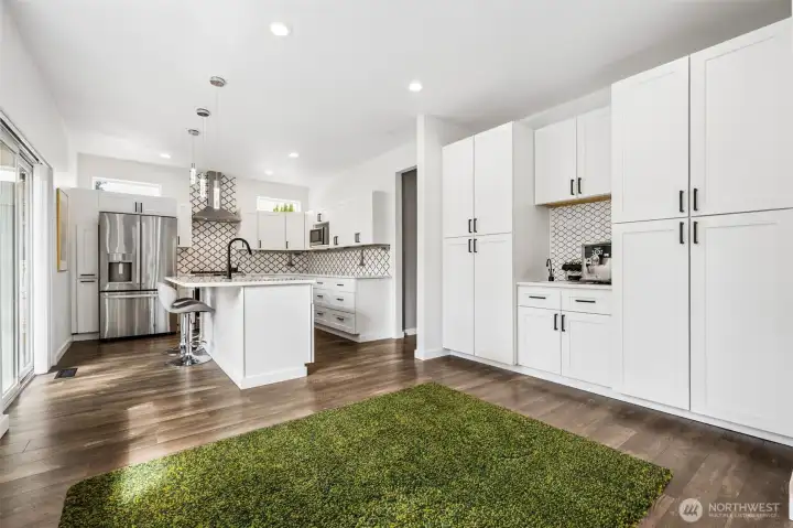 Crisp white cabinetry stretches floor to ceiling, creating a full feature wall that includes a secondary sink for a proper beverage or bar station. The island anchors the space with quartz surfaces and seating, while the continuous hardwood flooring keeps the entire area feeling unified. There’s flexibility built in here too, space for casual dining or keep it open depending on your style. Light moves easily through the room, and the layout carries you directly into the bedroom wing.