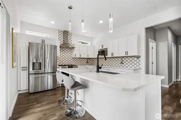 The island gives you front row seating for at least four, the kind of setup where conversations stretch longer than the meal. That French door fridge with the bottom freezer anchors the space, paired with a clean stainless vent hood that feels intentional, not builder basic. The microwave is built into the cabinetry where it belongs, out of your way but exactly where you need it. The fridge surround cabinetry is done right, seamless, elevated, no wasted space. And just when you think you’ve seen it all, those long horizontal windows above the cabinets pull in this soft, natural light that changes the whole mood. The hallway off to the right leads to the bedrooms, but this kitchen is absolutely the main event.