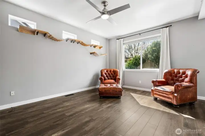 A large picture window brings in natural light, while those upper transom-style windows along the wall solve the usual layout problem, light pours in, privacy stays intact, and your furniture goes wherever you want it. Hardwood floors run through with that same warm, multi-tone grain, paired with a soft silver-gray wall color and crisp white trim that keeps everything sharp. A modern ceiling fan anchors the space overhead, and at roughly 12 by 16, there’s real room to spread out.
