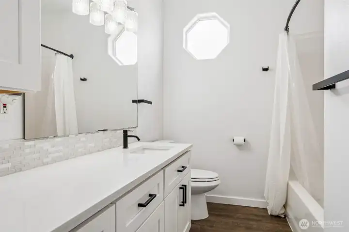 That long stretch of quartz gives you all the room in the world, no elbow battles here, paired with a crisp white glass tile backsplash that keeps everything feeling bright and fresh. The upper cabinet steps in with extra storage right where you want it, because clutter is not invited. The full tub and shower combo is clean and polished, ready for both quick mornings and long resets. And then that high octagon window, such a good moment, bringing in natural light while keeping everything completely private. Add in the oversized mirror, sleek sconces, matte black hardware, and sharp white cabinetry, and suddenly a “main bathroom” feels a lot more elevated than expected.