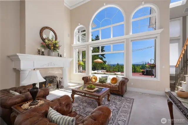 Sunlight pours through large windows, highlighting sweeping hilltop and mountain views. A glowing fireplace and striking staircase create a warm, inviting focal point.