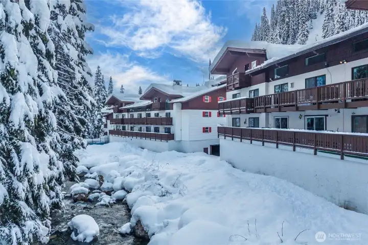 Charming alpine-style condo buildings set along a scenic river, surrounded by snow-covered evergreens and mountain views—capturing the true year-round beauty of Alpental.
