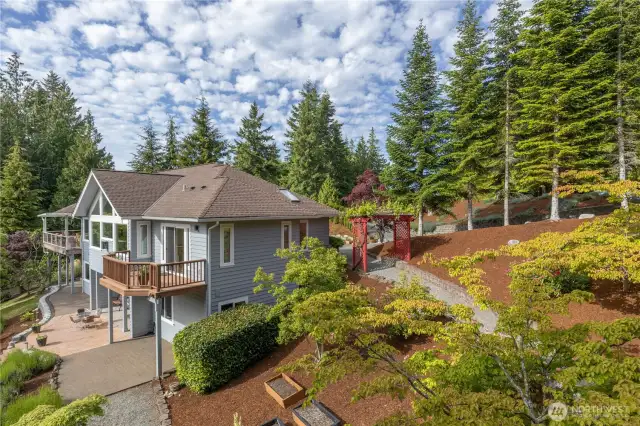 The south side of the home, showing the lovely gardens, rhododendrons. The deck in the forefront is off the primary bedroom, the covered deck in the background is off the kitchen. A hot tub pad exists on the lower patio.