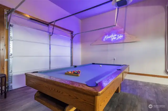 Garage game room with custom neon sign, professional grade pool table, ping pong, foosball, LVP flooring throughout, vehicle access at front and back of the garage.