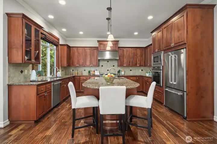 The expansive center island takes center stage in this chef's kitchen, offering abundant prep space and casual seating surrounded by rich cabinetry, gleaming granite countertops, and top-of-the-line stainless steel appliances. Elegant pendant lighting and acacia hardwood floors complete this beautifully appointed kitchen designed for both function and style.