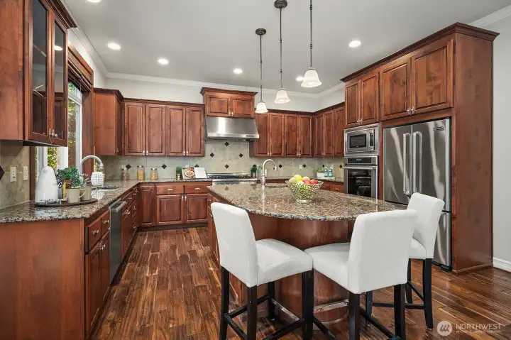 This sweeping view of the gourmet kitchen captures the full scope of the space, showcasing the abundant cabinetry, expansive granite countertops, and premium stainless steel appliances that make this kitchen a true entertainer's dream. Warm acacia hardwoods, elegant pendant lighting, and generous prep space come together to create a kitchen that is as beautiful as it is functional.