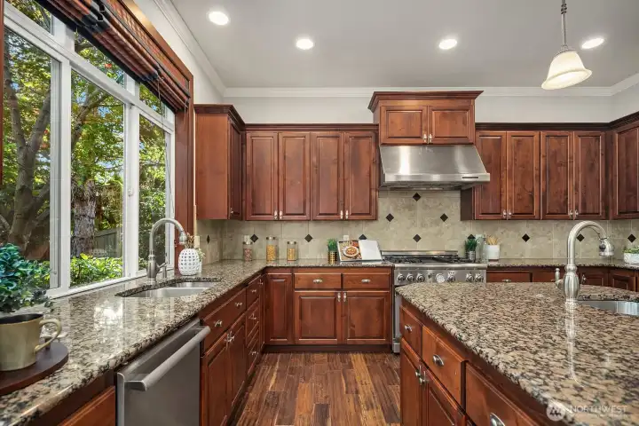 Natural light pours through the large window above the sink, creating a bright and cheerful workspace framed by rich cabinetry and a beautifully detailed tile backsplash. The generous granite countertops and premium stainless steel appliances, including a professional-grade range and hood, underscore the exceptional quality found throughout this gourmet kitchen.