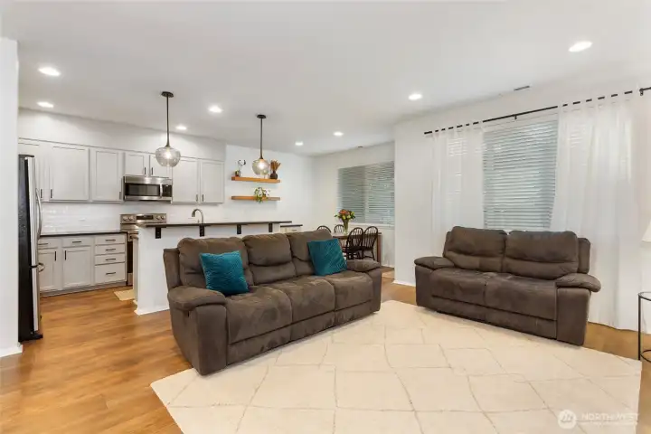 Bright open-concept living space featuring warm LVP floors, recessed lighting, and inviting natural light from oversized windows. The living room flows seamlessly into the upgraded kitchen, highlighted by a breakfast bar, modern pendant lights, and clean white cabinetry—ideal for both everyday living and entertaining.