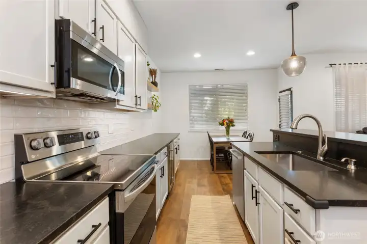 Another bright kitchen view highlighting the galley-style layout with granite countertops, stainless steel appliances, and modern cabinetry. Durable LVP flooring flows throughout, while recessed and pendant lighting enhance the clean, modern design and provide a clear sightline to the dining area—ideal for both everyday use and entertaining.