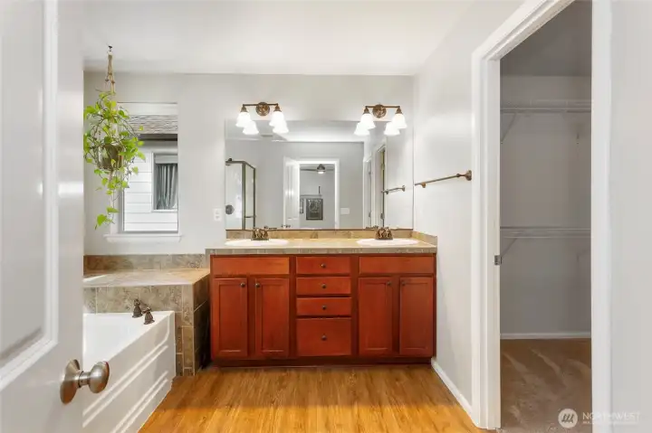 Primary bathroom with walk-in closet access featuring a spacious dual-sink vanity with warm wood cabinetry, generous counter space, and ample storage. Bright lighting, large mirror, and natural light create an open, airy feel. The door to the right leads to a massive walk-in closet, offering exceptional storage and a seamless, private flow within the primary suite.