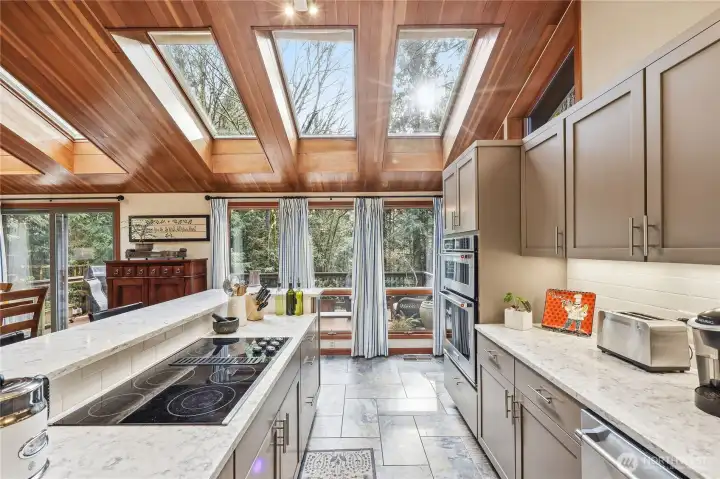 Another view of the kitchen looking toward the back deck with the cooktop on the left and the microwave, oven and dishwasher on the right. Note the ceramic tile floors throughout the kitchen space.