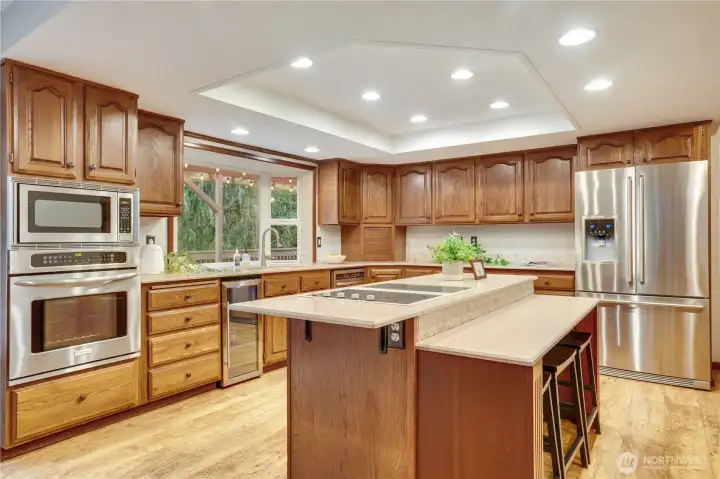 Spacious cook’s kitchen with plentiful cabinetry, center island with seating, stainless appliances, and a big window over the sink that looks out to the trees and deck.
