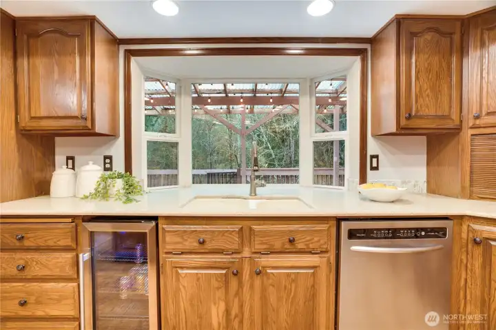 Sink-side prep zone with beverage fridge, tons of storage, and a big picture window over the sink that looks out to the covered deck and forested backdrop.
