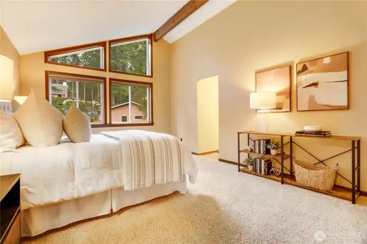 Serene primary bedroom with vaulted ceiling, exposed beam, and dramatic corner windows that frame the trees—creating a warm, retreat-like space to start and end your day.
