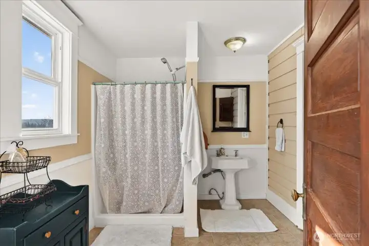 This classic 3/4 bathroom on the main floor is absolutely flooded with natural light from that big window. The white wainscoting keeps it feeling bright and airy. It has that authentic farmhouse charm, complete with a classic pedestal sink and a functional shower setup tucked into the corner. It is a perfectly quaint and cheerful space that stays true to the home's style.