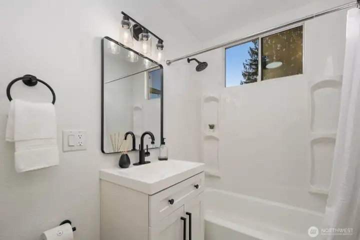 Updated full bath with a white vanity, black fixtures, framed mirror, and shower-tub combination with built-in shelving.