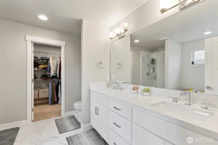 Large walk-in closet in the background.  Double vanity with quartz countertops and large wall mirror and beautiful floors.