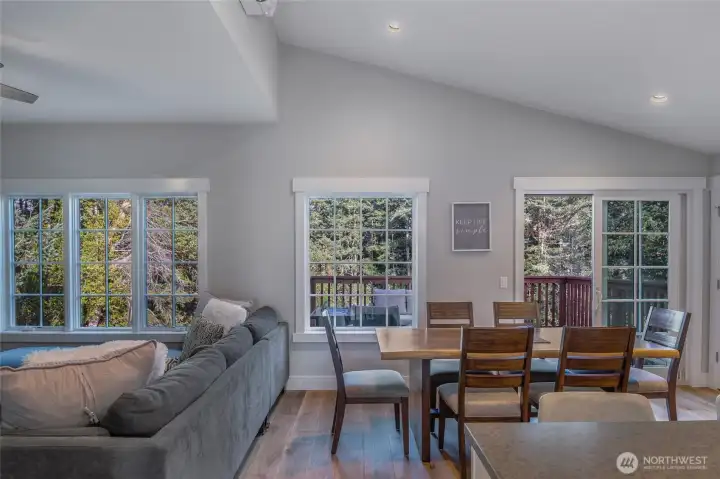 Light-filled open-concept living and dining area with vaulted ceilings, engineered wood floors, and large windows framing tranquil forest views—creating a bright, inviting space for everyday living in this Friday Harbor condo.