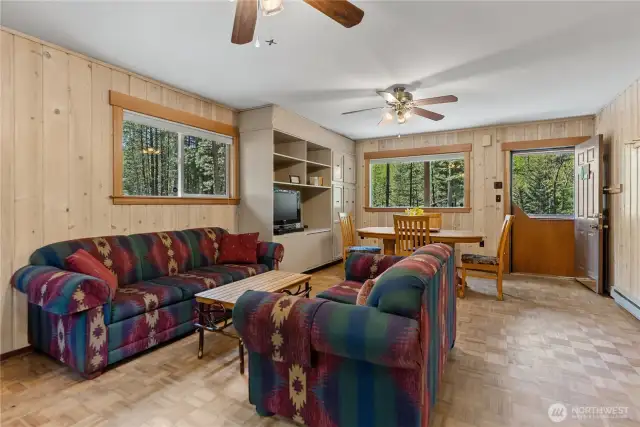 Warm up by the wood stove in this inviting space featuring parquet floors, knotty pine walls, and large windows framing forest and river scenes—classic cabin vibes with vintage flair.