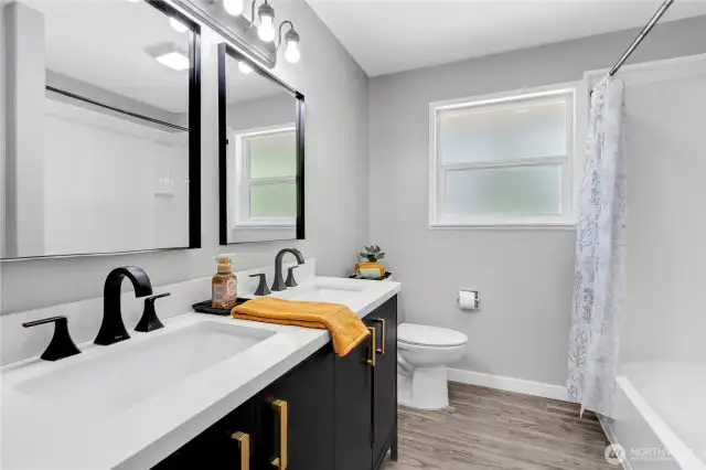 A beautifully renovated bathroom featuring a double vanity with sleek black fixtures, framed mirrors, and stylish gold hardware. The full-size tub and shower combo with a privacy-glass window complete the spa-like experience.