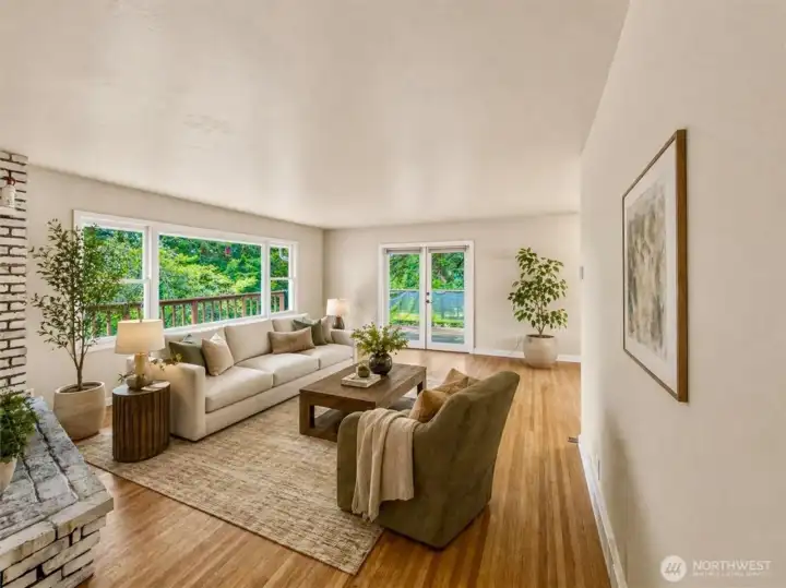 Large living room with abundant natural light, hardwood floors, and a wood-burning fireplace, with French doors leading to the wraparound deck. [virtually staged]