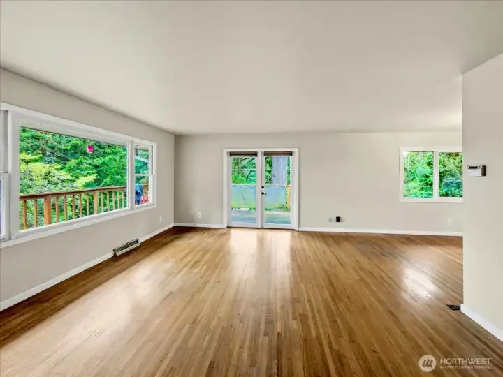 Large living room with abundant natural light, hardwood floors, and a wood-burning fireplace, with French doors leading to the wraparound deck. Additional dining area to the right.