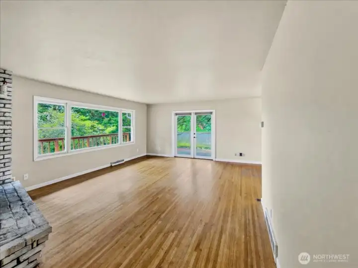 Large living room with abundant natural light, hardwood floors, and a wood-burning fireplace, with French doors leading to the wraparound deck. Additional dining area to the right.