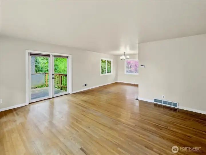 Large living room with abundant natural light, hardwood floors, and a wood-burning fireplace, with French doors leading to the wraparound deck. Additional dining area to the right.