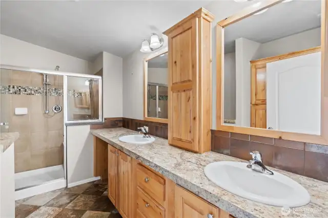 Beautifully upgraded primary bathroom featuring dual sinks, natural wood cabinetry, and a spacious walk-in shower with stylish tile accents. This luxurious ensuite adds both comfort and convenience to this energy-efficient home in Lacey’s 55+ Lakeview Meadows community.