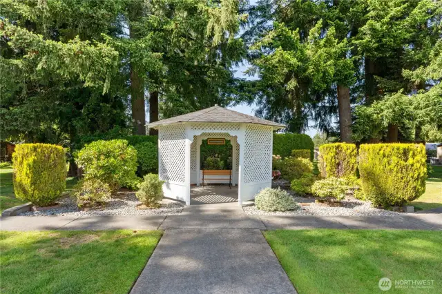 Charming community gazebo surrounded by lush landscaping and mature trees—perfect for peaceful strolls or quiet moments outdoors. This serene green space is one of many inviting features in the 55+ Lakeview Meadows neighborhood in Lacey, WA.