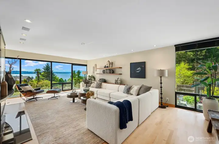 Light-filled living room with expansive picture windows showcasing stunning water views. Open, airy layout features hardwood floors, recessed lighting, and a modern fireplace