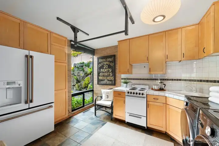 Light-filled secondary kitchen with warm wood cabinetry, tiled surfaces, and garden views