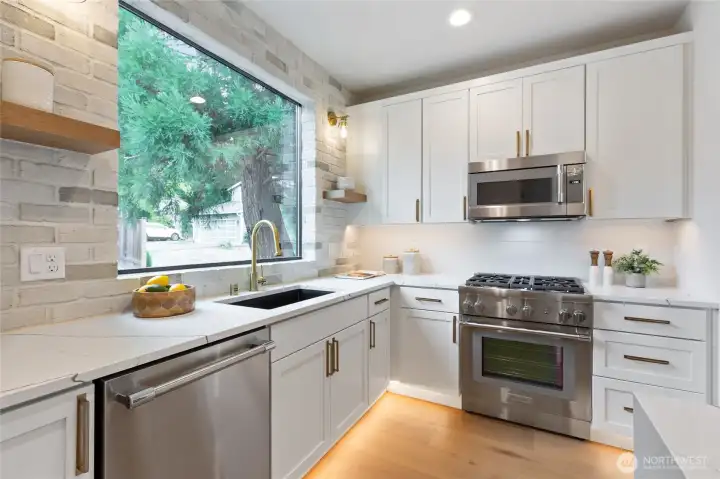 This stylish kitchen pairs crisp white cabinetry and gold hardware with sleek stainless-steel appliances and a soft gray brick backsplash—elevated by a large window that floods the space with natural light and warmth.