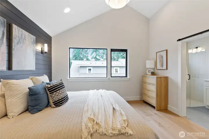 This bedroom offers a cozy, modern retreat with layered textures, a dark paneled accent wall, and vaulted ceilings—brightened by natural light and softened with warm wood tones.
