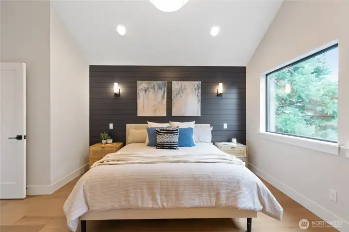 This spacious bedroom features a vaulted ceiling, dark paneled accent wall, and soft layered textures—framed by a large window that fills the room with natural light and treetop views.