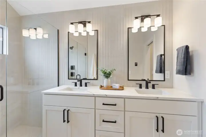 This modern bathroom features a double vanity with black-framed mirrors and sleek hardware, balanced by a glass-enclosed shower and soft natural accents for a clean, symmetrical look.