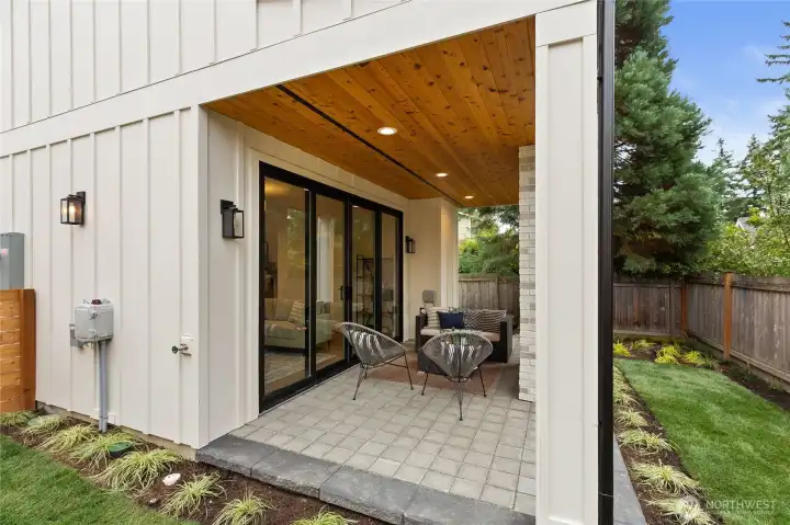This stylish covered patio blends indoor comfort with outdoor charm, featuring recessed lighting, and a cozy seating area—all framed by lush landscaping and easy access through sliding glass doors.