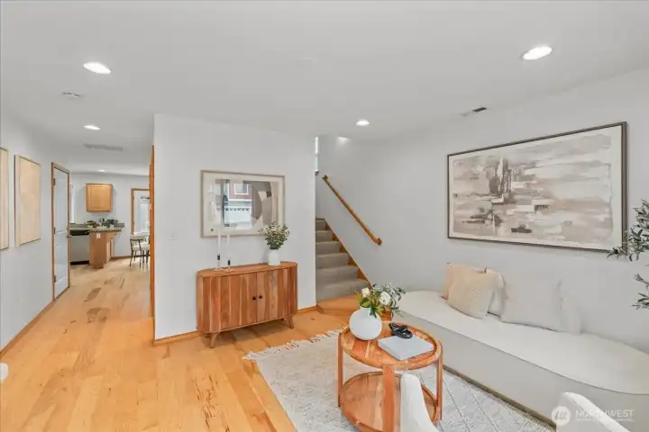 Light-filled living area near the front entry featuring laminate flooring, recessed lighting, and a large front-facing window. Open layout connects the living space to the hallway and kitchen area.