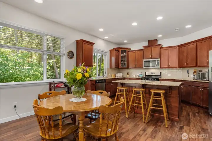 Bright kitchen and dining area featuring warm wood cabinetry, granite counters, island seating, large windows, and views of the surrounding evergreens. Bright kitchen and dining area featuring warm wood cabinetry, granite counters, island seating, large windows, and views of the surrounding evergreens.