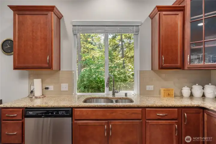 Kitchen sink area with granite countertops, tile backsplash, warm cabinetry, stainless dishwasher, and peaceful wooded views from the window. Kitchen sink area with granite countertops, tile backsplash, warm cabinetry, stainless dishwasher, and peaceful wooded views from the window.