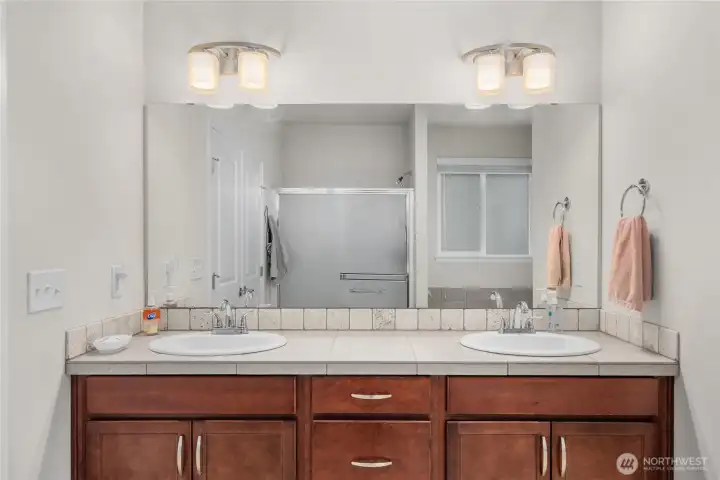 Dual-sink primary bathroom vanity with wood cabinetry, tile countertop, large mirror, and updated light fixtures. Dual-sink primary bathroom vanity with wood cabinetry, tile countertop, large mirror, and updated light fixtures.
