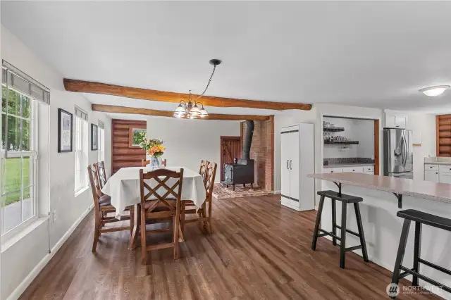 Open Dining & Kitchen Layout with Wood Stove Warmth  This open-concept space brings together the dining area, kitchen & cozy wood stove nook—offering functionality, warmth & charm in one flowing layout. The exposed log beams, wood-style flooring & natural light from multiple windows create an inviting environment for family gatherings or entertaining. The breakfast bar adds extra seating & convenience, while the classic wood stove provides a rustic focal point with true Pacific Northwest character.