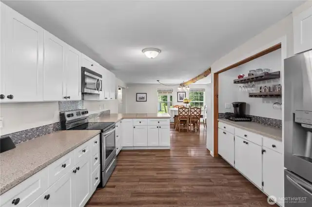 Modern Farmhouse Kitchen with Stainless Steel Appliances  This beautifully updated kitchen offers the perfect balance of rustic charm & modern functionality. Featuring sleek stainless steel appliances, crisp white cabinetry, solid-surface countertops & a stunning glass tile backsplash, this space is ideal for everyday cooking or hosting guests. The rich wood flooring adds warmth, while the windowed log wall brings in natural light & a touch of that cozy Pacific Northwest cabin character. A dream kitchen for any country home enthusiast!