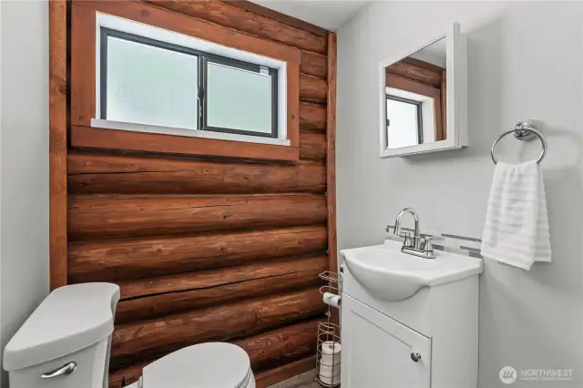 Rustic-Modern Half Bath with Log Wall Accent  This charming half bath blends modern touches with rustic appeal, featuring a beautiful log wall accent that brings warm cabin character to the space. A sleek white vanity, updated fixtures & mosaic backsplash add a clean, fresh feel, while the frosted window offers natural light with privacy. Perfectly placed for guests or daily convenience, this bathroom adds style & function to your Pacific Northwest retreat.