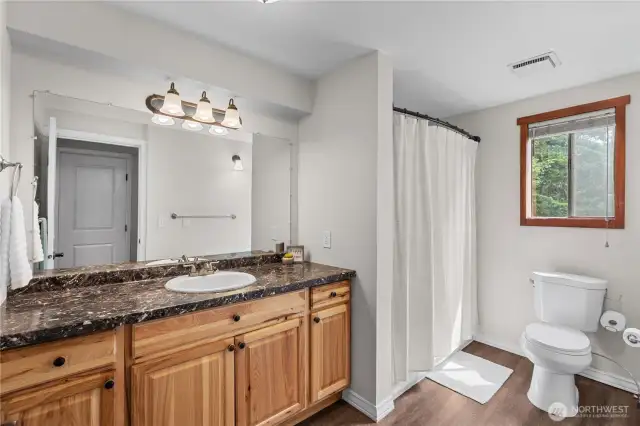 Full Bathroom with Rich Wood Cabinetry & Natural Light  This spacious full bath features beautiful natural wood cabinets, a dark stone-style countertop, & a wide mirror with triple-light vanity—delivering both warmth & functionality. The shower/tub combo with updated fixtures offers everyday convenience, while the large window brings in serene views & soft natural light. A perfect blend of rustic charm & modern comfort, ideal for guests or everyday use in this Pacific Northwest retreat.