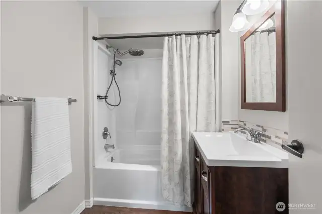 Charming Upper-Level Bathroom  Fresh, functional, and full of style—this upstairs bathroom features a full-size tub with a rainfall showerhead, sleek tile accents, and a modern vanity with rich wood cabinetry. Perfectly placed for easy access to all upstairs bedrooms, it's both beautiful and practical.
