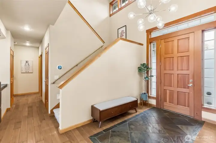 Right when you walk in, you can tell this home has presence. The eight panel solid wood door is framed with wood wrapped trim and flanked by tall privacy windows, plus that long accent window above that brings in light without putting your life on display. The slate detail at the entry handles wet shoes like a pro, then transitions into those rich maple floors that run through the main level. The space opens up to a full two story foyer with a clean, modern chandelier that keeps it feeling current. There’s room for a bench setup, which just makes everyday comings and goings easier. You’ve got quick access to the powder room, a hallway leading to the main floor suite, and a large under stair storage area that’s way more useful than expected.