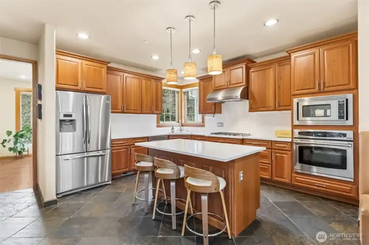 This view leans right into the kitchen’s “I’ve got this” energy. The quartz island is bright and inviting, surrounded by solid cherry cabinets that bring a richer, more custom feel than your standard setup. The backsplash runs full height in large white tiles, which keeps everything looking crisp and cohesive. Cooking here is actually enjoyable with the five burner cooktop, plus the separate wall oven and microwave setup that frees up space where you need it. A brand new 5 month old stainless dishwasher is already in place, and the French door fridge with water, ice, and a pull out freezer checks all the boxes. Overhead, three sconces light the island perfectly, making it just as good for late night snacks as it is for full dinner prep.