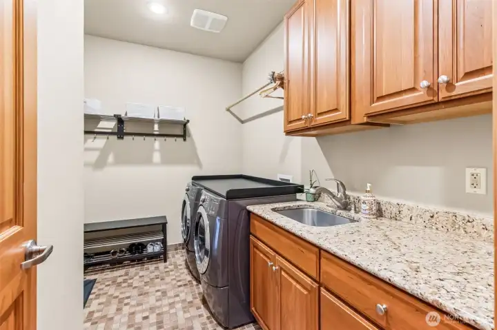 This is the kind of laundry room that makes everyday routines feel a little more organized. It’s on the main floor, spacious, well laid out, and gives you room to actually work. The long granite counter stretch all the way to the machines and there is even a solid surface on top creating the perfect folding, sorting, or staging laundry baskets area. The washer and dryer stay and already have a countertop insert in place, which just makes life easier. An undermount sink adds flexibility, while upper cabinetry and that hanging rod keep things functional. Tile floors handle the wear and tear, and being right off the garage, it doubles as a smart drop zone that keeps the rest of the house a little more put together.